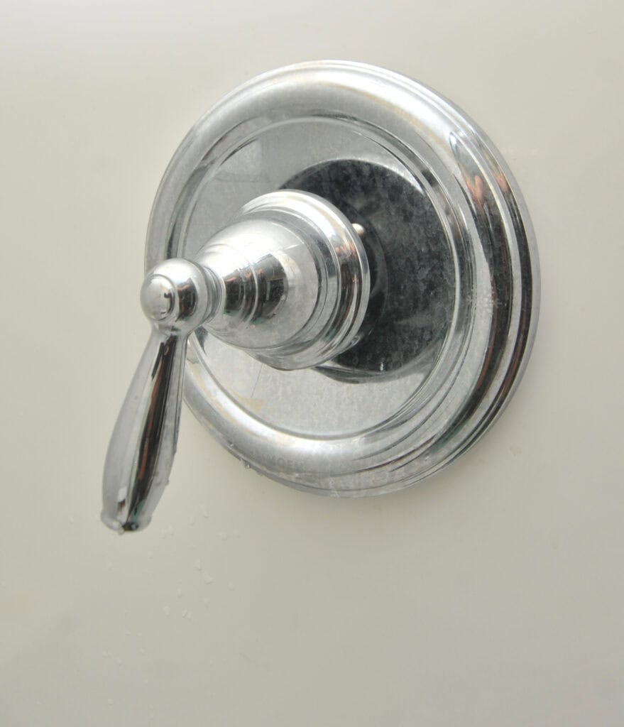 A close-up of a shiny metal shower or bathtub faucet handle mounted on a white wall, with water droplets visible on the surface.