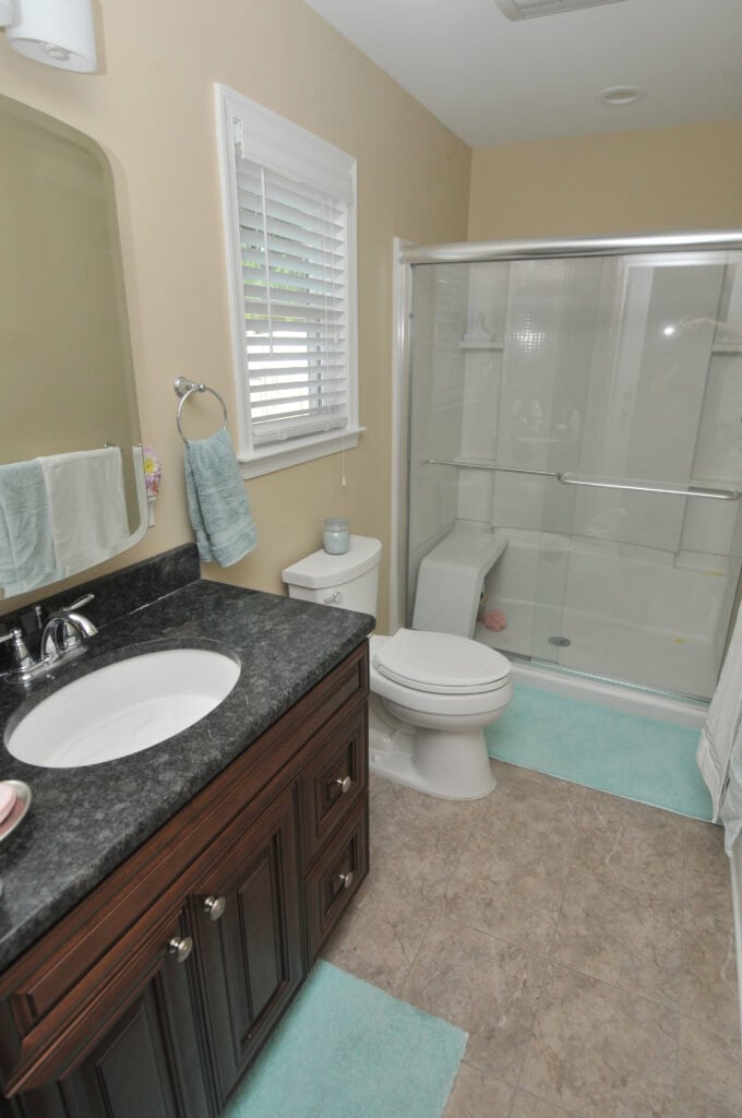 A bathroom with a dark wood vanity, black countertop, sink, mirror, white toilet, shower with sliding glass doors, and light blue bath mats on tiled floor. Light beige walls and a window with white blinds.
