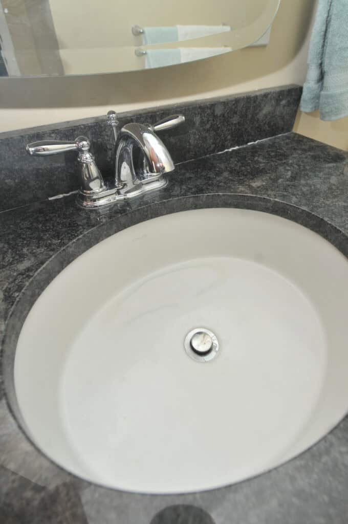 A white oval bathroom sink with a silver faucet set in a dark marble countertop. A mirror and a towel are visible in the background.