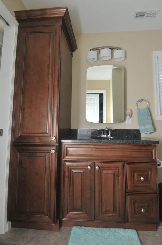 A bathroom vanity with dark wood cabinets and drawers, a black countertop, a mounted rectangular mirror, three-light fixture above, silver faucet, towel ring with teal towel, and a teal bathmat on the floor.