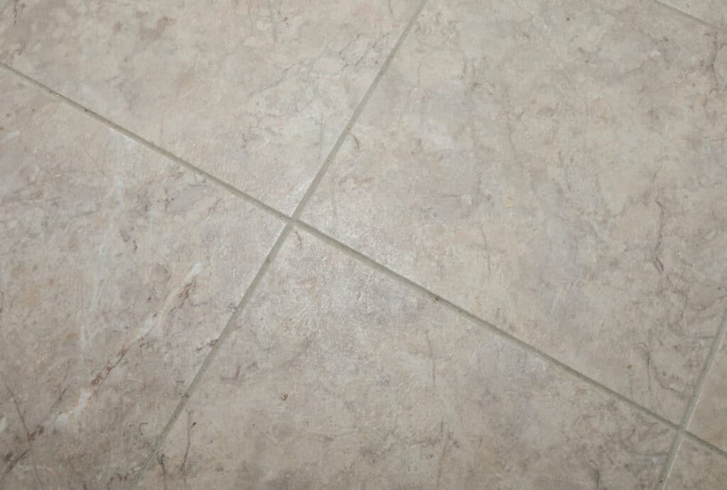 Close-up of beige ceramic floor tiles with light gray grout lines forming a grid pattern; the tiles have a subtle marbled texture.