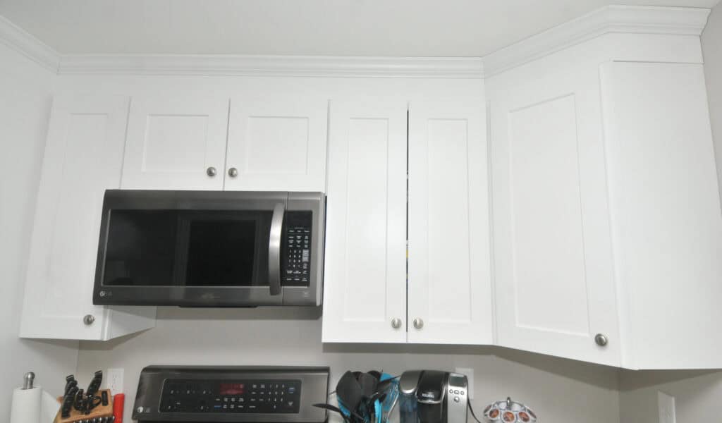 White kitchen cabinets with silver knobs are mounted above a stainless steel microwave and stove. Small kitchen appliances and utensils are on the counter below. The cabinets have a simple, modern design.