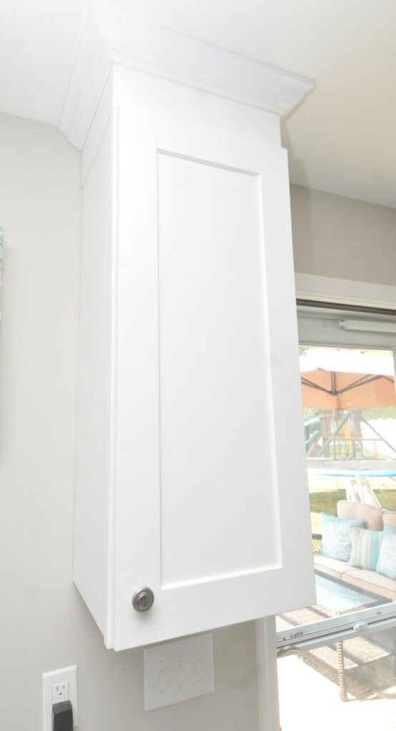A tall, white, rectangular kitchen cabinet with a simple panel door and silver knob is mounted on a beige wall next to a window with a view of an outdoor patio.
