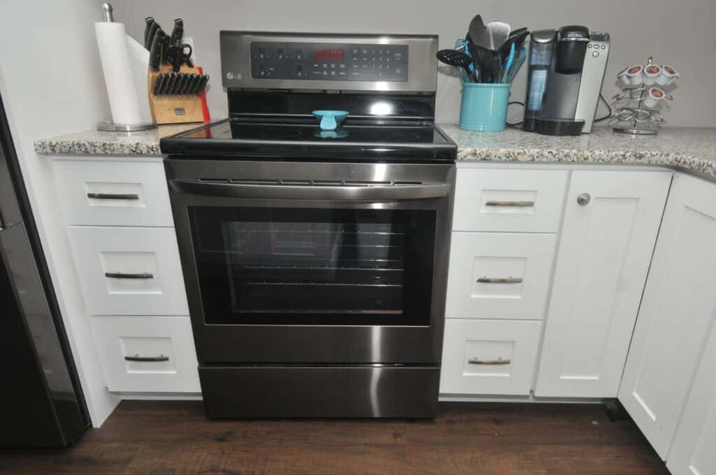 A modern kitchen featuring a stainless steel electric stove with a blue handle on the oven door, white cabinets, a granite countertop, utensils in a teal holder, a Keurig coffee maker, and a knife block.