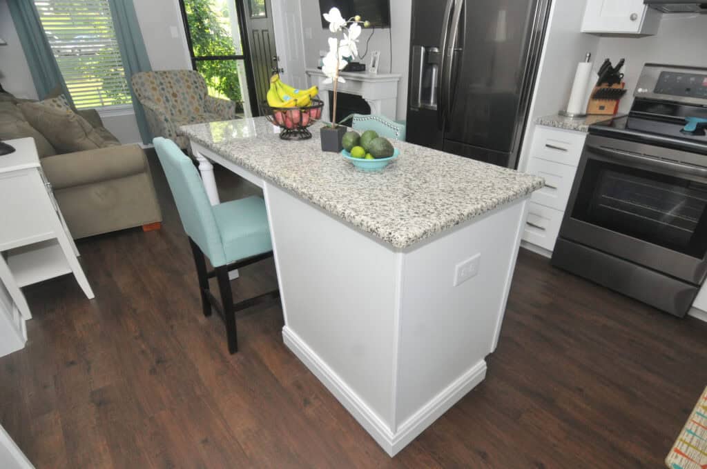 A modern kitchen with a granite island, teal chairs, a bowl of bananas, a fruit basket, and a bowl of limes. Stainless steel appliances and dark wood floors are visible, with a sofa and window in the background.