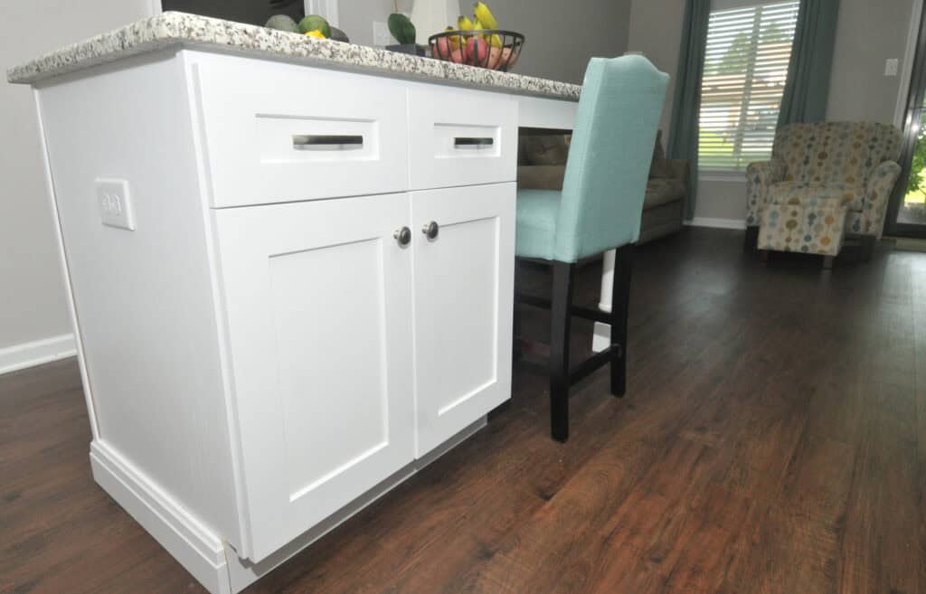 White kitchen island with granite countertop, turquoise upholstered barstool, hardwood floors, and a fruit bowl on top. A patterned armchair and window with blinds are visible in the background.