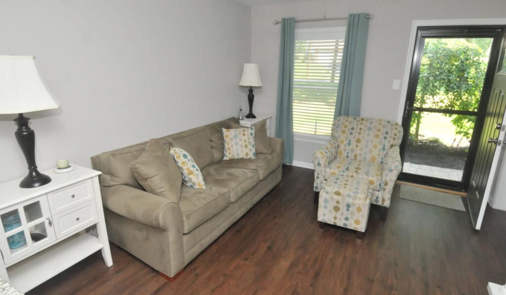 A small living room with a beige sofa, patterned armchair, two lamps, a white side table, wood floor, and a window with blue curtains. The front door is open, showing greenery outside.