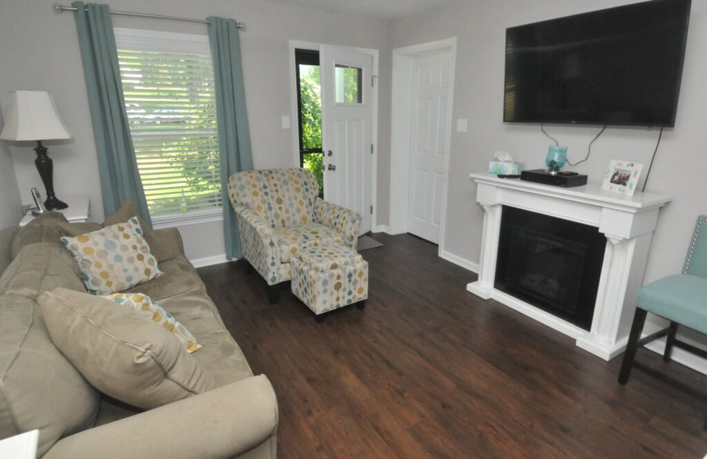 A cozy living room with a beige sofa, patterned armchair, teal curtains, a fireplace with a TV mounted above, and wooden floors. Light streams in through a window beside the front door.