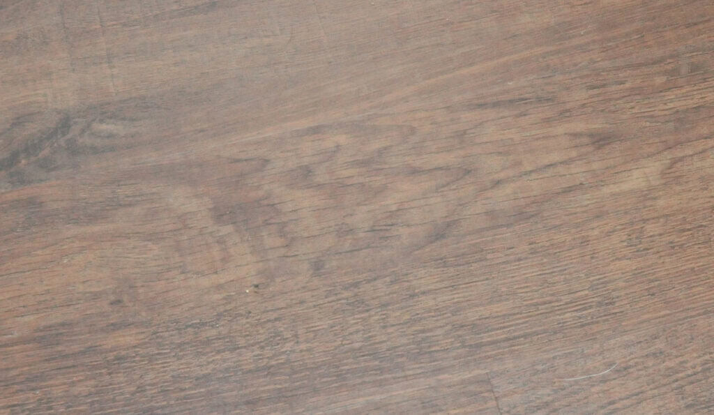Close-up of a wooden surface with visible grain patterns, natural knots, and varying shades of brown. The wood texture appears slightly worn and rustic.