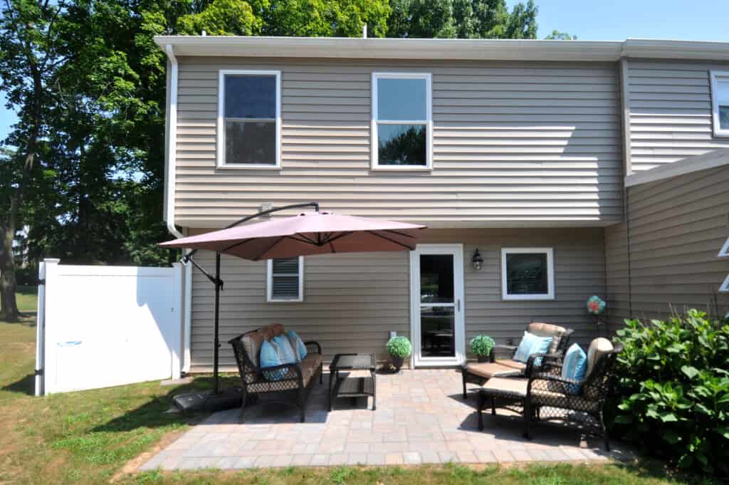 A backyard patio with wicker furniture, cushions, a coffee table, a large umbrella for shade, and potted plants, set against a beige house with two stories and a white fence on a sunny day.