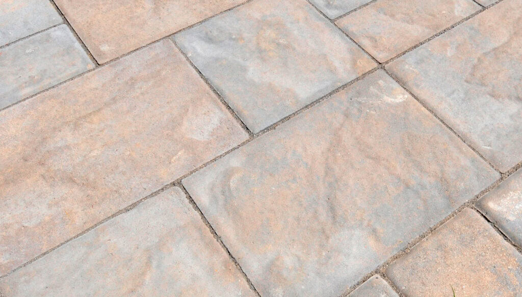 Close-up of a paved surface with large rectangular stone tiles in shades of beige and gray, arranged in a staggered pattern. The texture appears slightly rough and weathered.