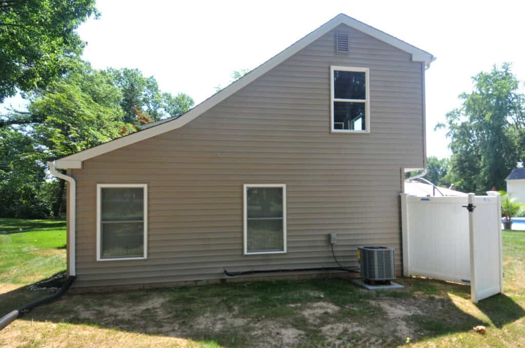A beige house with three rectangular windows, tan siding, and a sloped roof, next to a white fence and outdoor air conditioning unit, with green trees and grass in the background.
