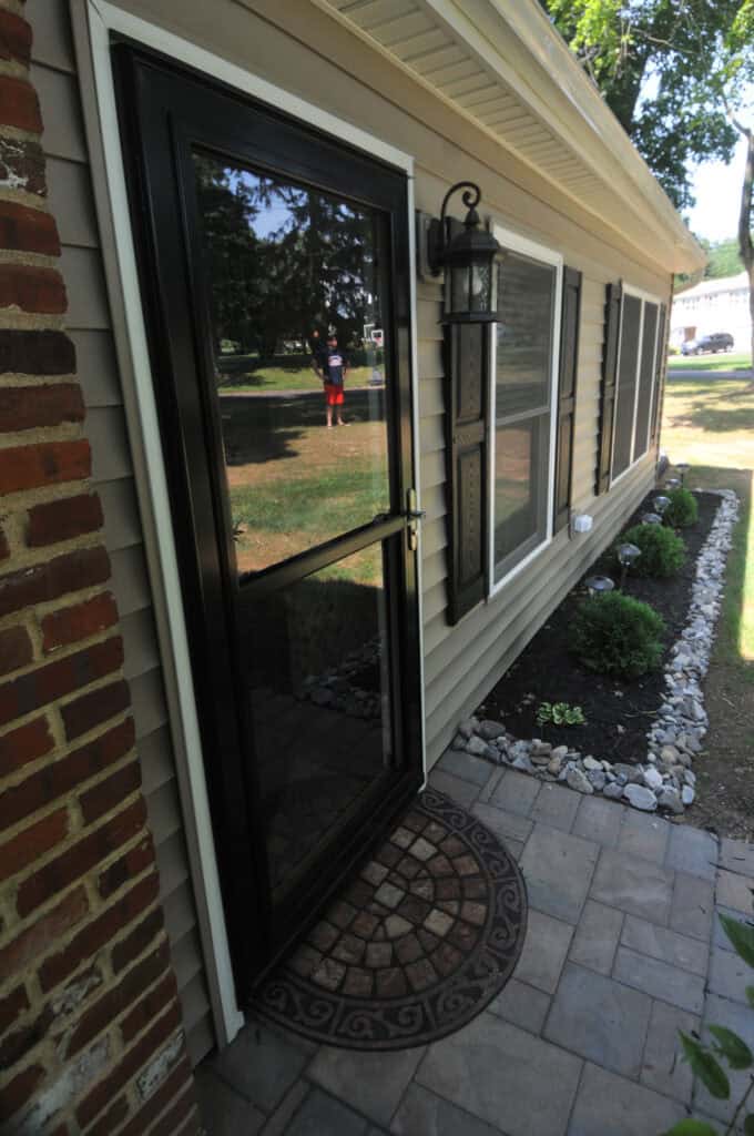 A glass storm door with a black frame is open, revealing the entrance to a tan house with white trim. There is a round decorative doormat and a small landscaped garden with white stones along the side.