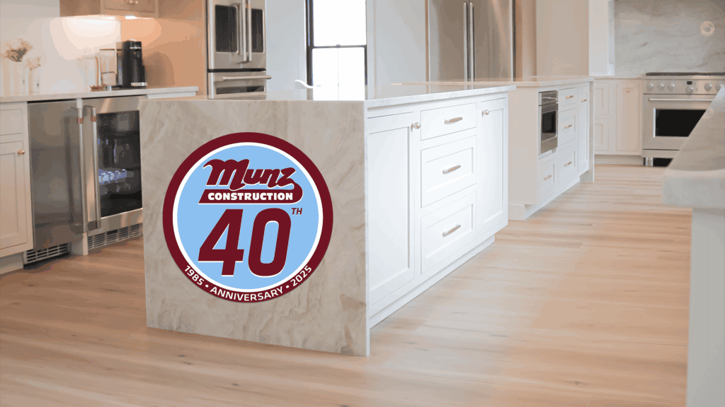 A modern white kitchen with light wood floors. The end of the island displays a large Munz Construction 40th anniversary logo, dated 1985–2025—a subtle reminder to consider what should I know before remodeling my home.