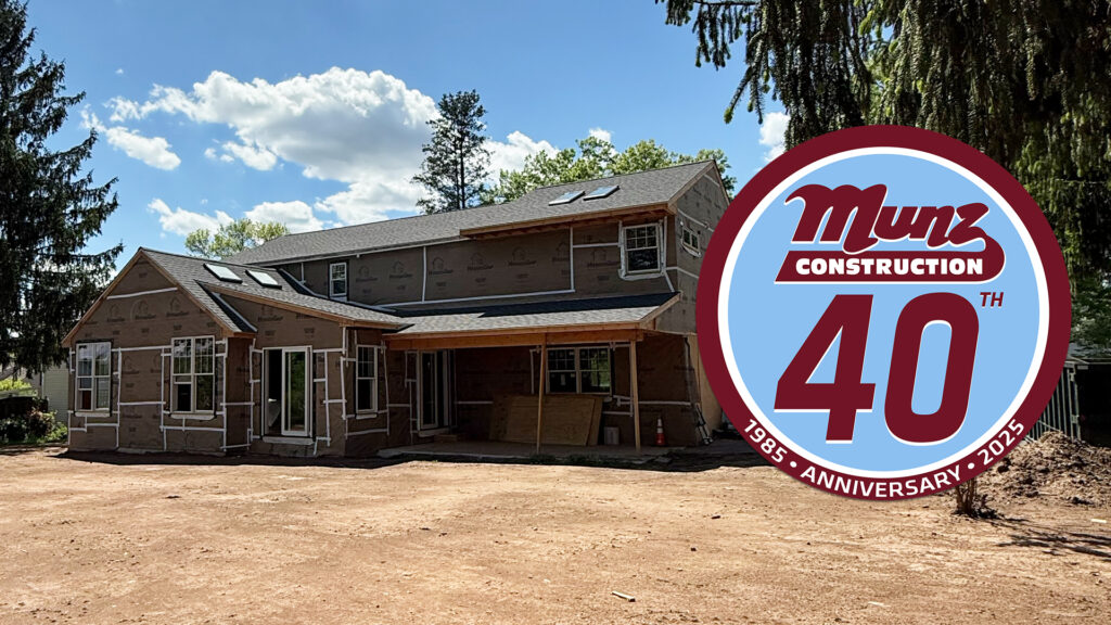 A large two-story house under construction, surrounded by dirt and trees, highlights the importance of asking what should I know before remodeling my home, with a circular Munz Construction 40th Anniversary 1985-2025 logo overlayed on the right.