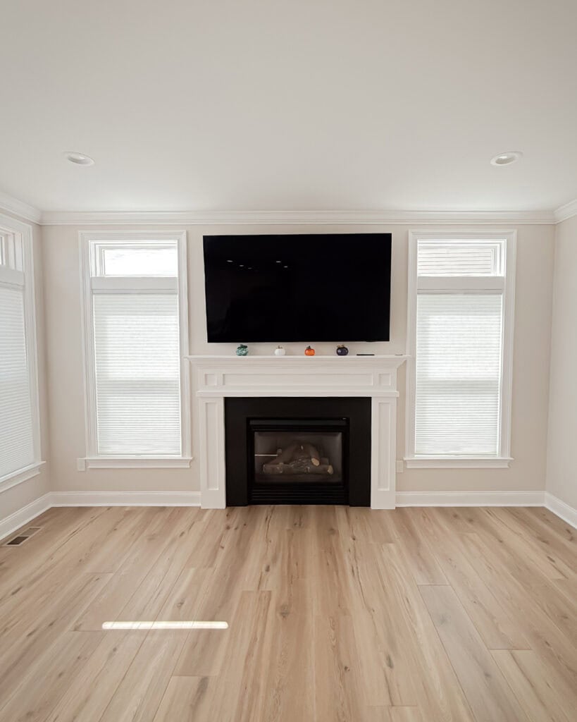 A modern living room with light wood floors, a white fireplace, a black flat-screen TV mounted above it, and two tall windows with blinds on either side of the fireplace.