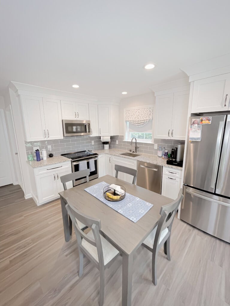 Modern kitchen with white cabinets, stainless steel appliances, gray subway tile backsplash, and light wood flooring. A light wood dining table with four chairs sits in the center, decorated with a blue runner and a fruit bowl.