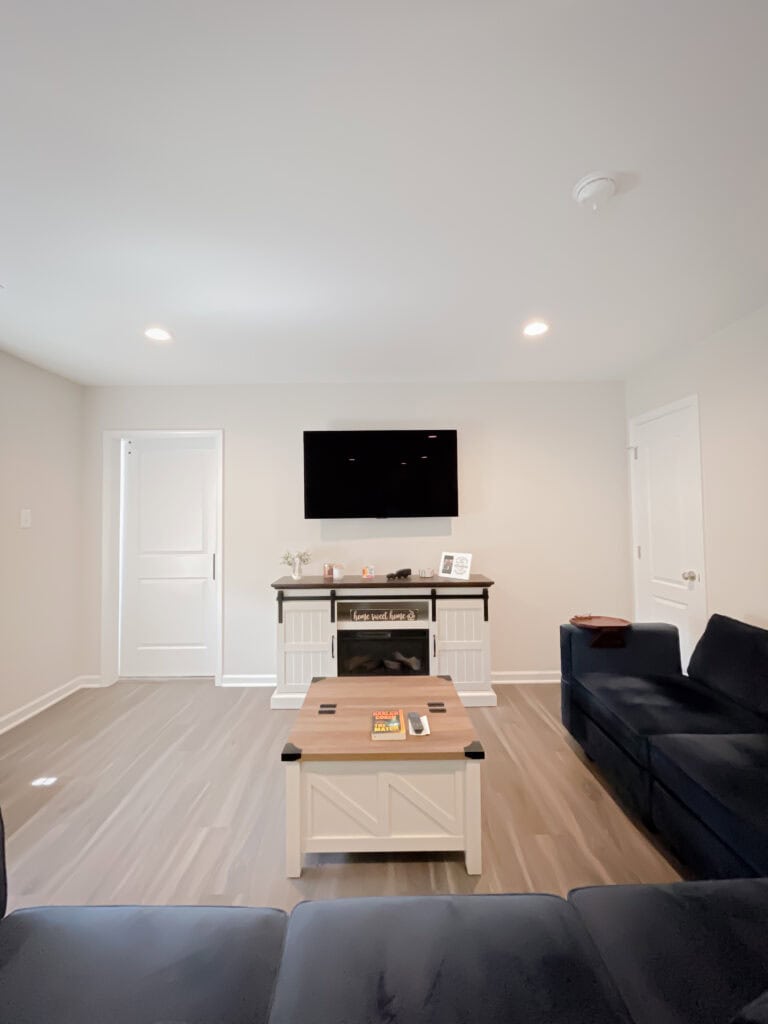 A modern living room with light-colored walls and floors, a black sofa, a white coffee table, and an entertainment console with a TV mounted on the wall above it. Two closed doors flank the console.