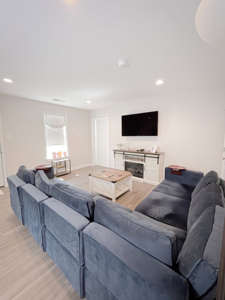 A bright living room with a large blue sectional sofa, a white coffee table, a small side table, a TV mounted above a white fireplace, and light-colored wood flooring. A window lets in natural light.