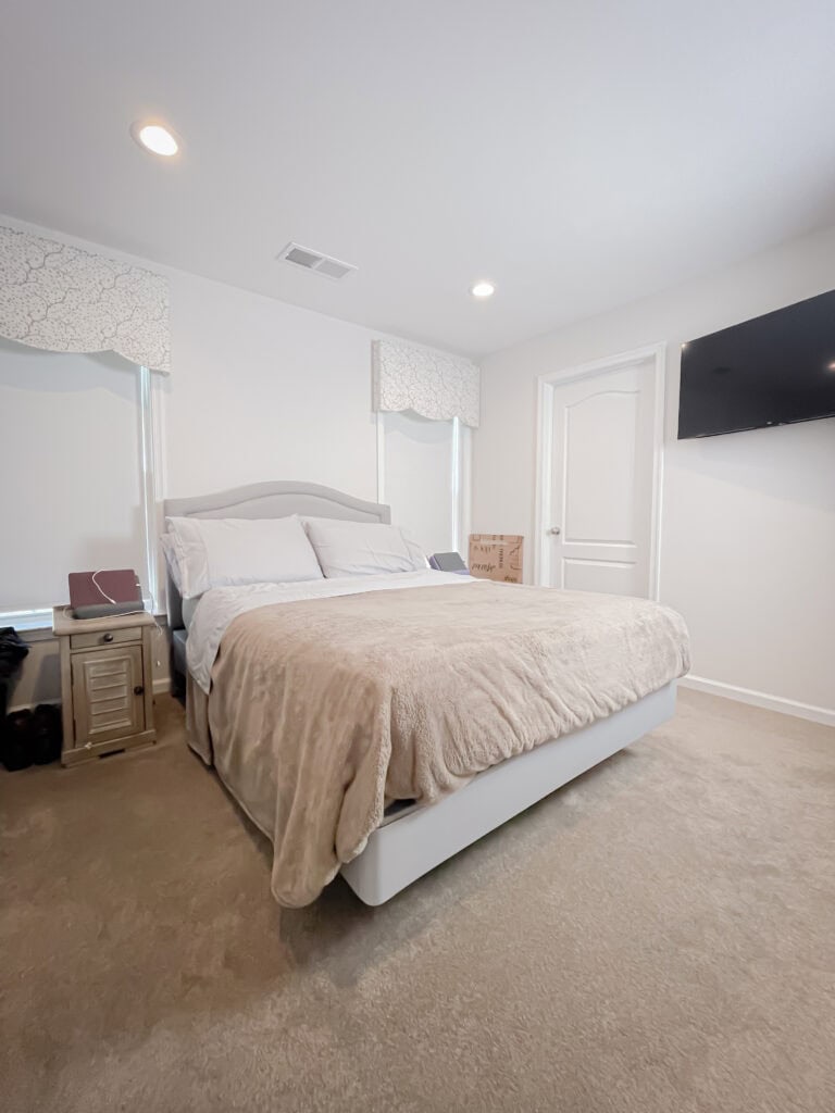 A bright, modern bedroom with a neatly made bed covered in a tan blanket, two nightstands, a wall-mounted TV, and beige carpet. The room has white walls, ceiling lights, and a box on the far side of the bed.