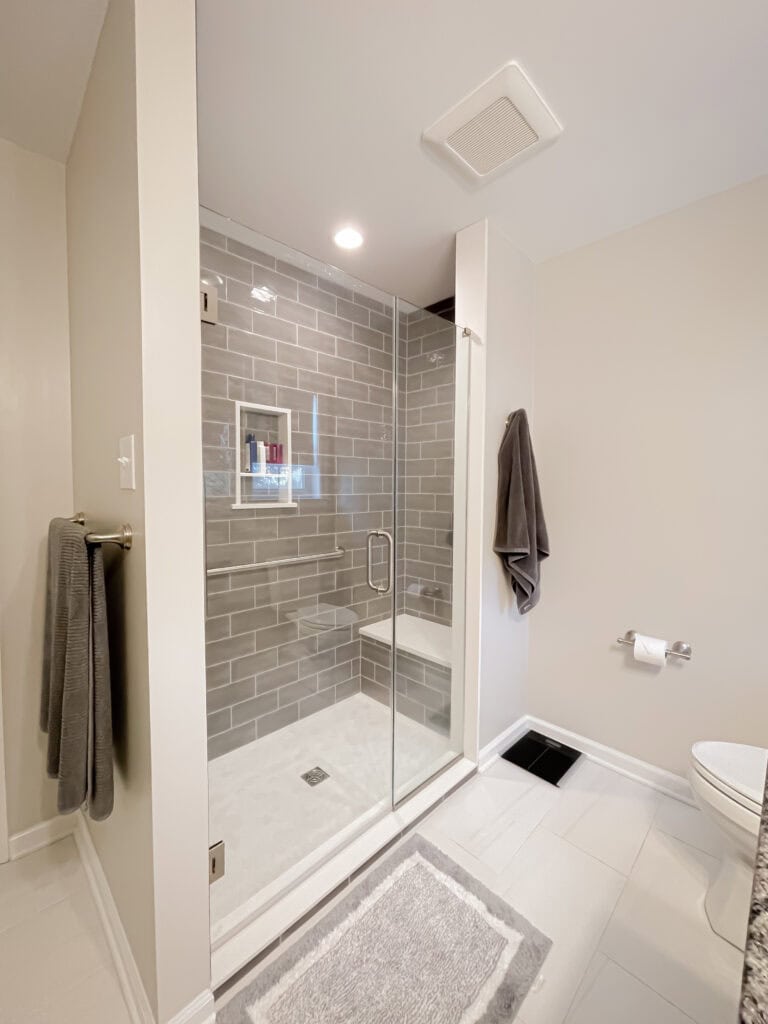 Modern bathroom with a glass-enclosed shower featuring gray subway tiles, a gray towel on a wall hook, a matching towel on a rack, a toilet, and a gray and white rug on a white tile floor.