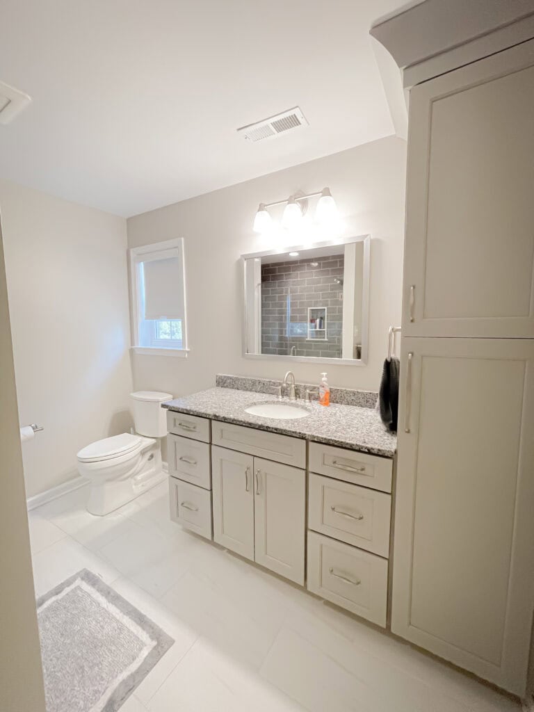 Modern bathroom with light gray cabinets, granite countertop, rectangular mirror with lights above, white toilet, towel, soap dispenser, window, and a gray rug on white tile floor. Walls are painted a soft beige.