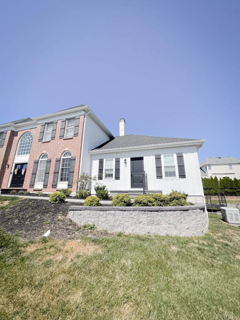 A white single-story house with black shutters and a black door sits next to a two-story red brick house, both under a clear blue sky with a green lawn in front.
