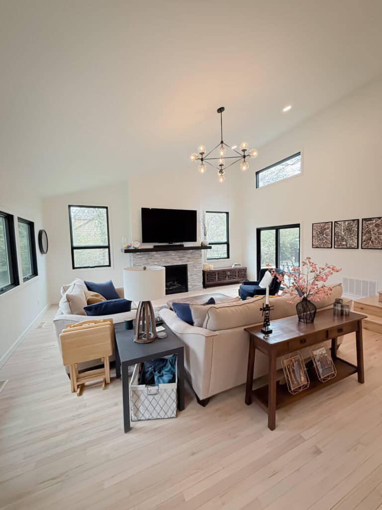 Modern living room with light wood floors, cream walls, and large windows. Features a beige sectional sofa, TV above a fireplace, a console table with decor items, and a stylish chandelier overhead.