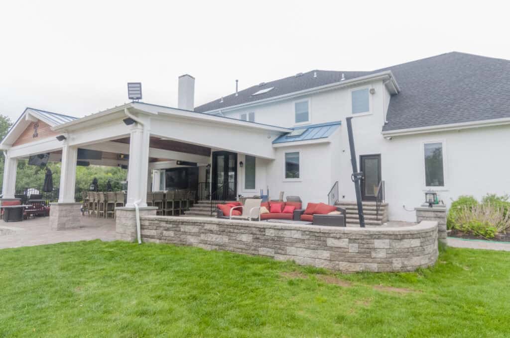 A modern two-story white house with a covered patio, outdoor dining area, stone retaining wall, and red cushioned patio furniture, overlooking a well-kept lawn.