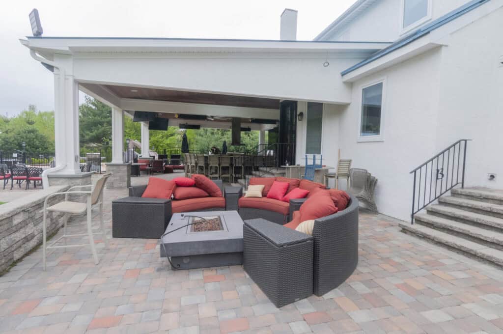 Outdoor patio with wicker furniture, red and orange cushions, and a fire pit table on a stone-tiled surface. Covered area with bar seating and greenery in the background. Steps lead to a white house.