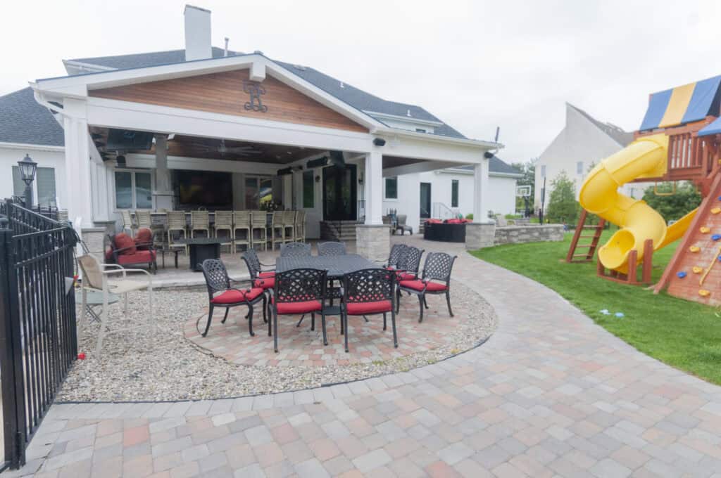 A backyard patio with a table and red-cushioned chairs, an outdoor bar under a covered area, and a wooden playset with a yellow slide on the grass.