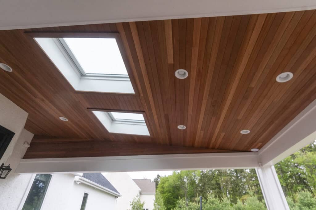 A wooden patio ceiling with built-in recessed lights and two rectangular skylights, allowing natural light to enter. White beams frame the ceiling, and trees and part of a house are visible outside.