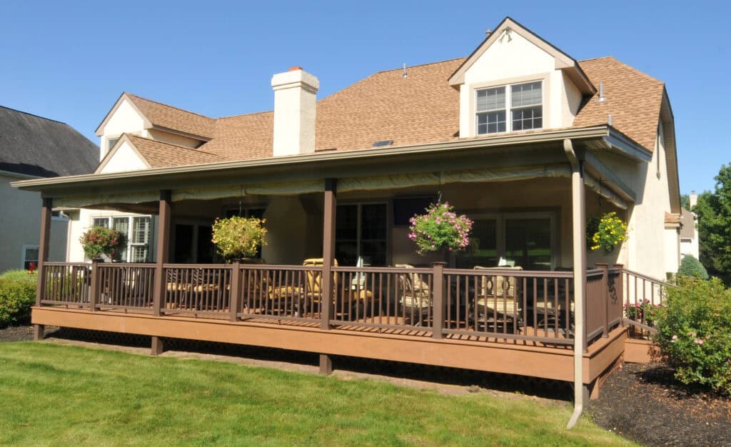 A beige house with a brown shingle roof has a covered wooden deck featuring railings, hanging flower baskets, chairs, and tables, surrounded by green grass and shrubs under a clear blue sky.