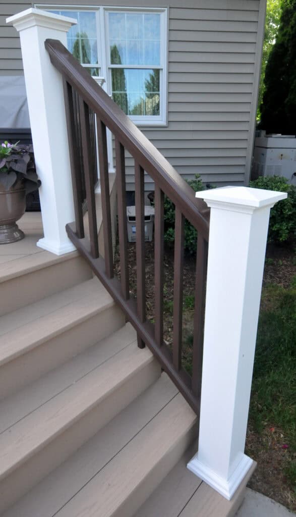 A beige outdoor staircase with white posts and a brown wooden handrail, leading up to a house with tan siding and a window reflecting greenery. A potted plant sits at the top of the steps.