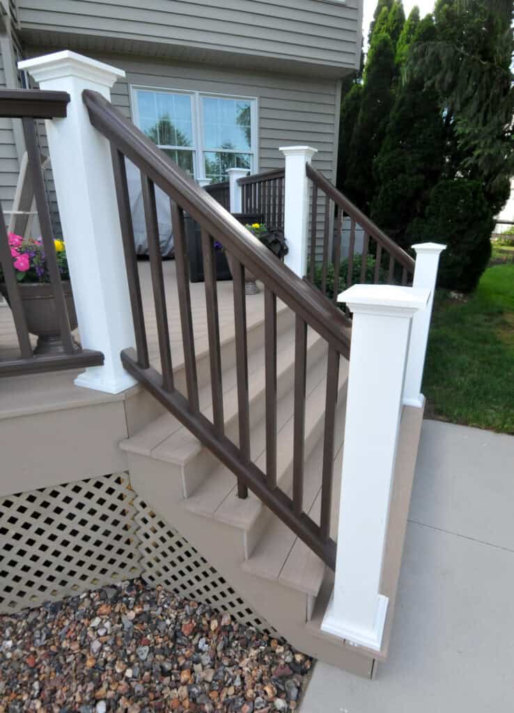 A small wooden staircase with brown steps, white and brown railing, and white posts leads up to a porch. There are plants and flowers in pots, rocks beside the steps, and evergreen shrubs in the background.