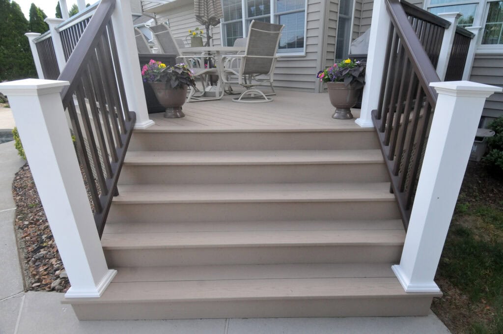 A set of five light brown porch steps with dark brown railings and white posts leads up to a deck with patio furniture and two potted plants on either side of the stairs.