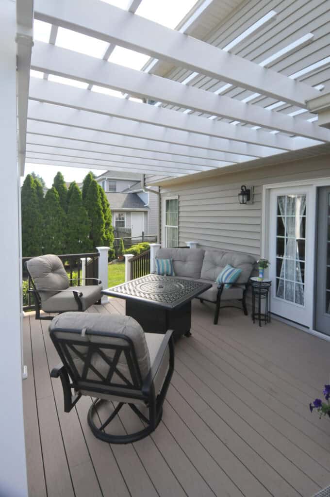 A cozy outdoor patio with beige cushioned chairs, a small table, and a pergola overhead, attached to a house with beige siding and French doors. Green bushes and neighboring houses are in the background.