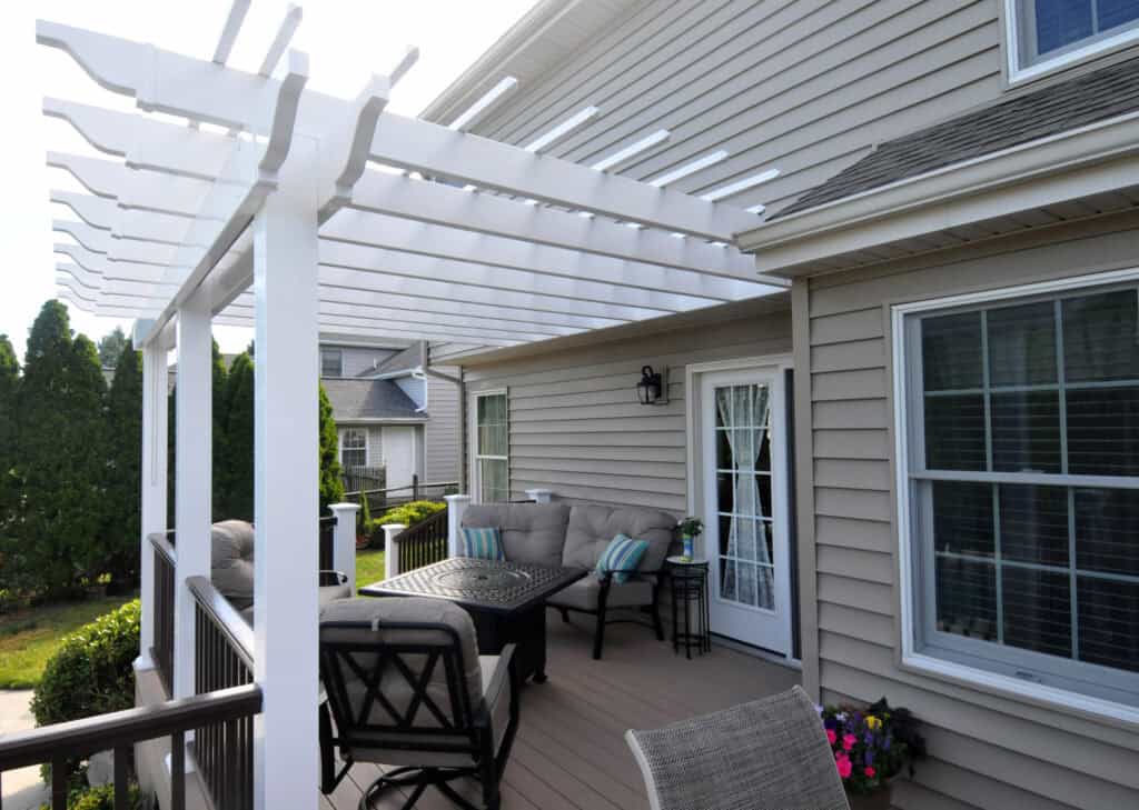 A backyard patio with beige siding, a white pergola, outdoor seating, a fire pit table, potted flowers, and a glass door leading into the house. Shrubs and neighboring homes are in the background.
