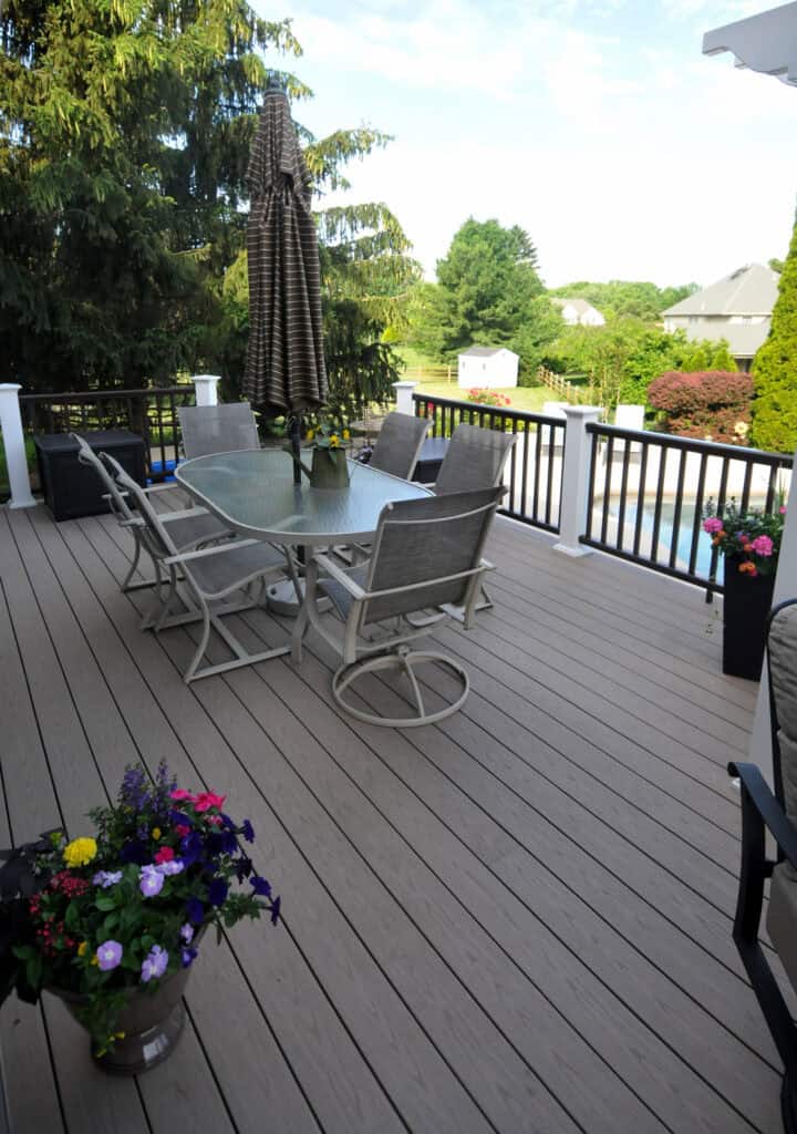 A spacious backyard deck with a glass table, six chairs, and a striped umbrella. Flower pots with colorful blooms sit on the deck, and a pool and greenery are visible in the background.