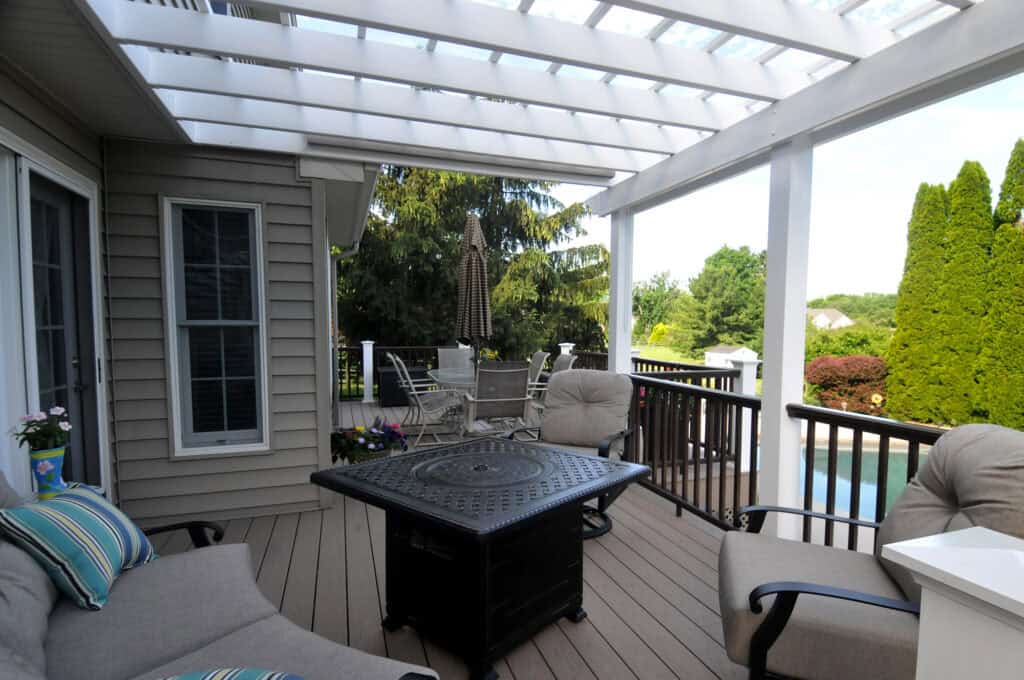 A cozy outdoor patio with cushioned chairs, a fire pit table, a dining set in the background, and a pergola overhead. The space overlooks a garden with trees and a glimpse of a swimming pool.