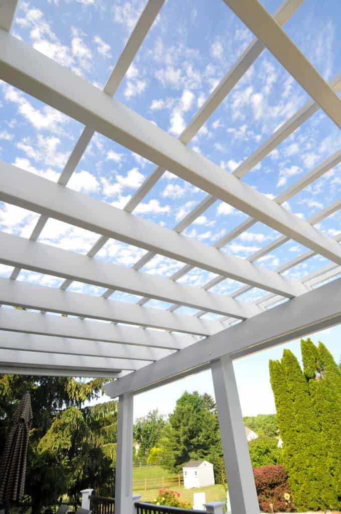 White pergola with rectangular beams viewed from below, set against a bright blue sky with scattered clouds. Green trees and a grassy yard are visible in the background.