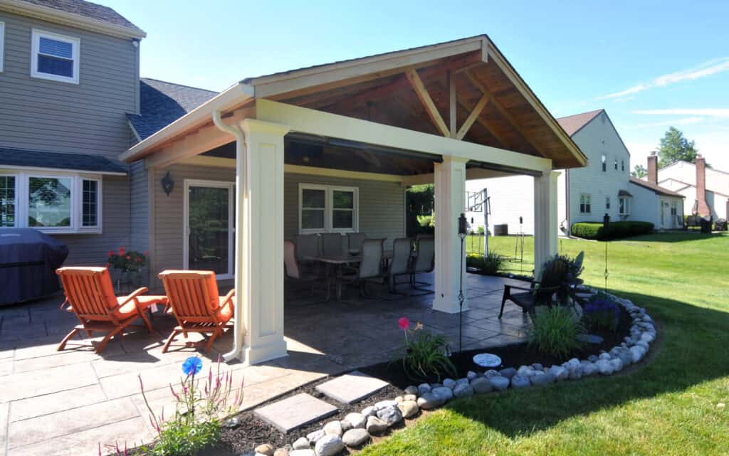 A covered patio with a dining table, chairs, and orange lounge chairs sits beside a two-story house. The patio overlooks a grassy yard, landscaped garden, and neighboring homes under a sunny sky.