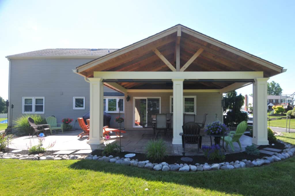 A backyard patio with a wooden pavilion, outdoor furniture including chairs and a table, surrounded by landscaping with rocks, flowers, and a grassy lawn. A two-story house is in the background.