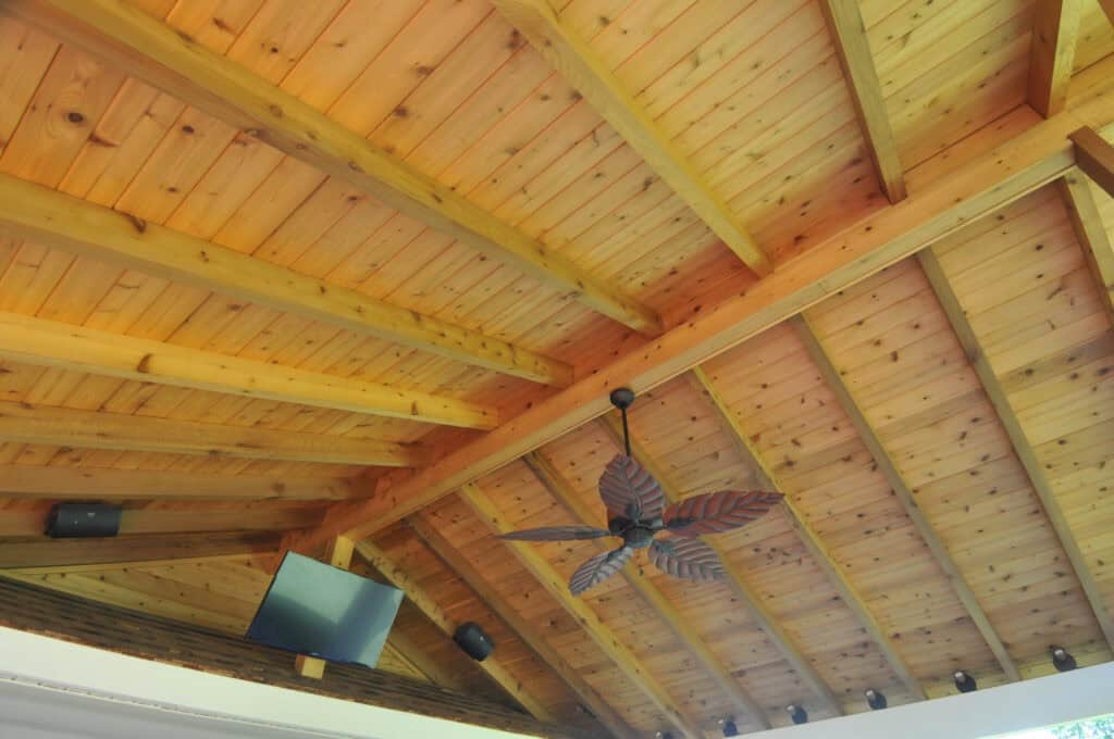A wooden ceiling with exposed beams, a leaf-shaped ceiling fan, several small black speakers, and a mounted flat-screen TV in the corner.
