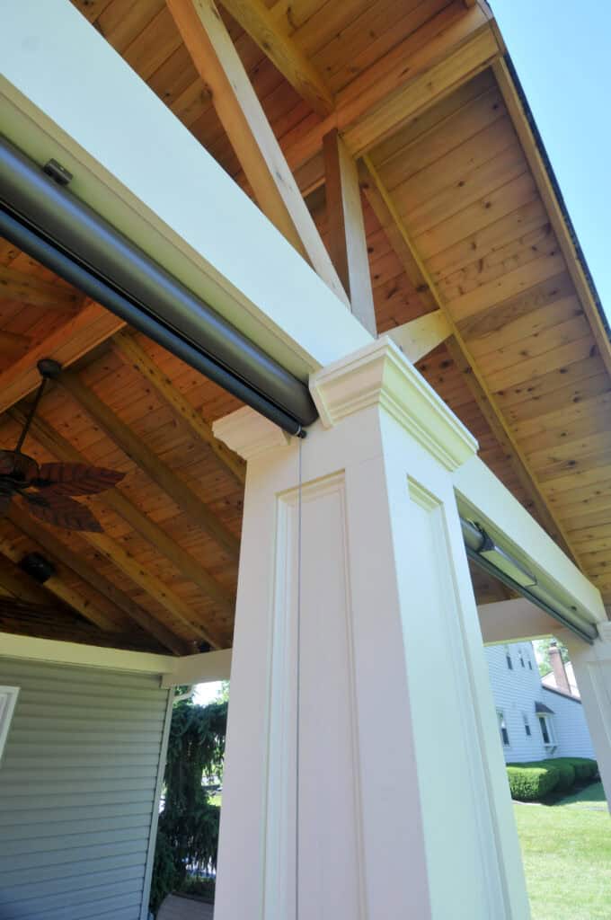 A close-up view of a white column supporting a wooden ceiling with exposed beams in an outdoor covered patio area, with the sky and neighboring houses visible in the background.