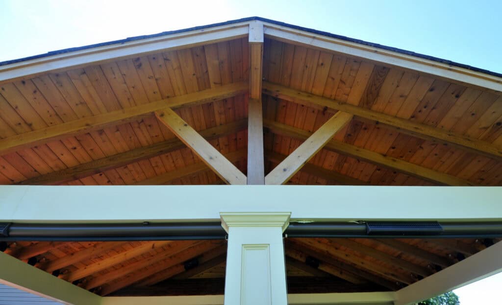 View looking up at the wooden ceiling and beams of a covered outdoor structure with a central white column supporting the roof against a clear blue sky.