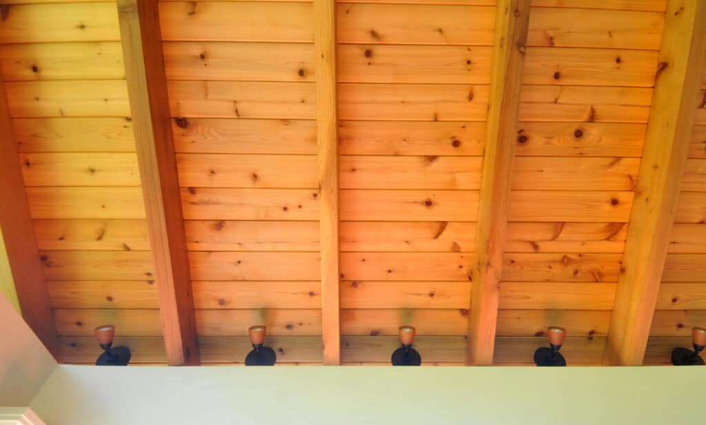 View of a wooden ceiling with visible beams and knots, featuring a row of small black and copper spotlights mounted along the base where the ceiling meets the wall.