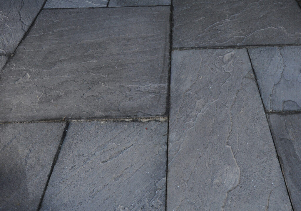 Close-up view of gray stone paving slabs arranged in a rectangular pattern, showing textured surfaces and slightly uneven grout lines between the stones.