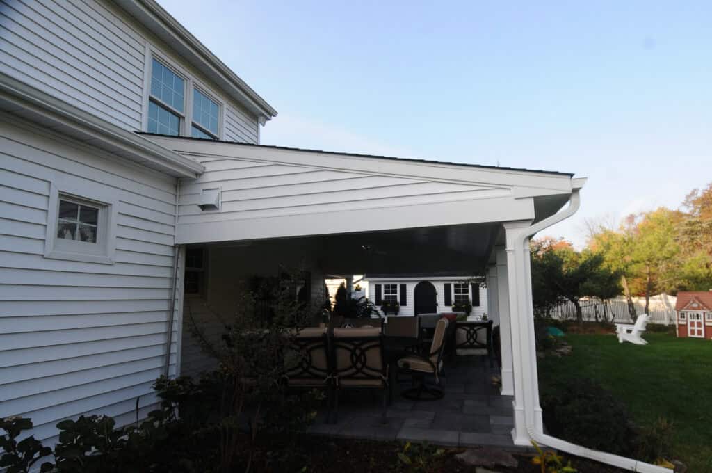 A covered patio attached to a white house with outdoor dining furniture underneath, next to a green lawn and trees, with a small red playhouse and garden shed in the background.
