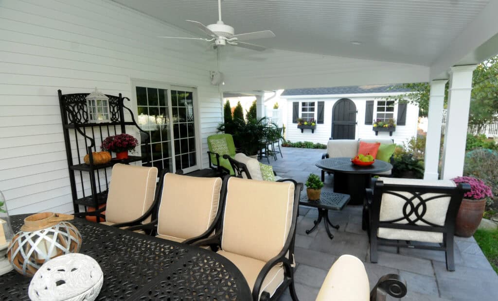A covered patio with cream cushioned chairs, black metal tables, potted plants, and flowers. In the background is a white garden shed with double doors and window boxes. The area appears clean and inviting.